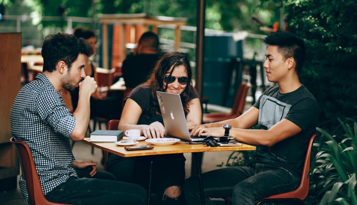 A group of young adults working on a laptop at an outdoor coffee shop, enjoying teamwork and collaboration.
