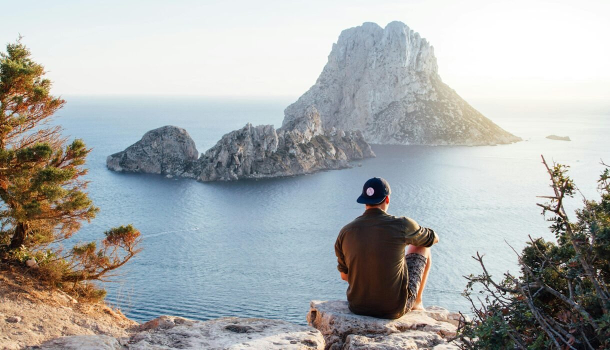 Man enjoys a scenic view of Es Vedrà at sunset from a cliff in San Juan Bautista, providing a perfect summer escape.