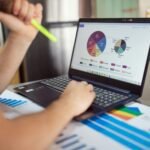 Person analyzing financial charts and graphs on a laptop with colorful documents, showcasing market analysis.
