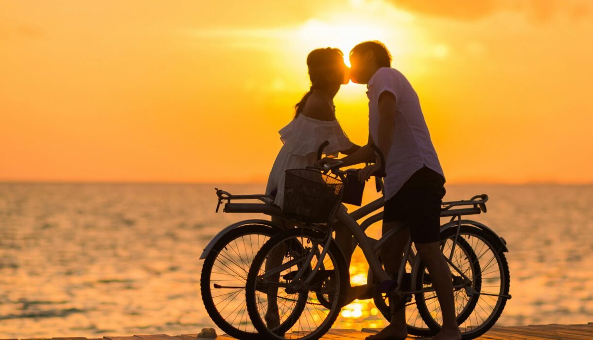A couple shares a romantic kiss on bicycles during a beautiful sunset at the beach.