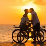 A couple shares a romantic kiss on bicycles during a beautiful sunset at the beach.
